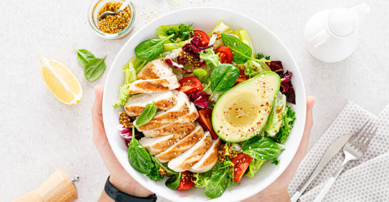 Grilled chicken meat and fresh vegetable salad of tomato, avocado, lettuce and spinach. Healthy and detox food concept. Ketogenic diet. Buddha bowl in hands on white background, top view